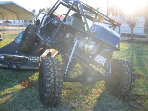 1992 Ford Mustang - A heavily modified off-road vehicle is precariously balanced on top of a dark sedan in a grassy yard.