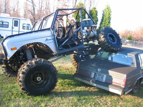 1992 Ford Mustang - A heavily modified off-road vehicle is perched atop a passenger car in a grassy area.