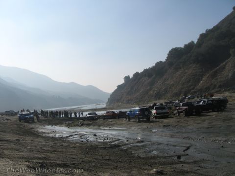 1987 Suzuki Samurai - A gathering of off-road vehicles and people are seen near a riverbed with a steep, vegetated hillside in the background.