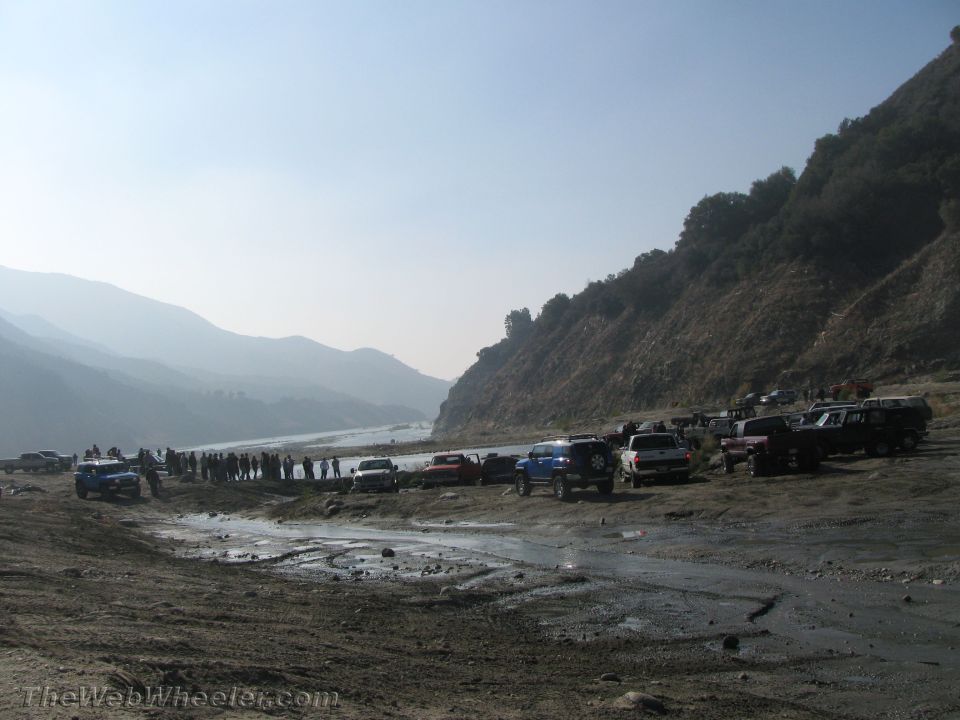 group of people and many off-road vehicles parked near a river and a large hill