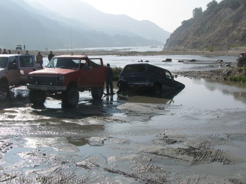 1987 Suzuki Samurai - Multiple off-road vehicles are stuck in a muddy riverbed with mountains in the background.