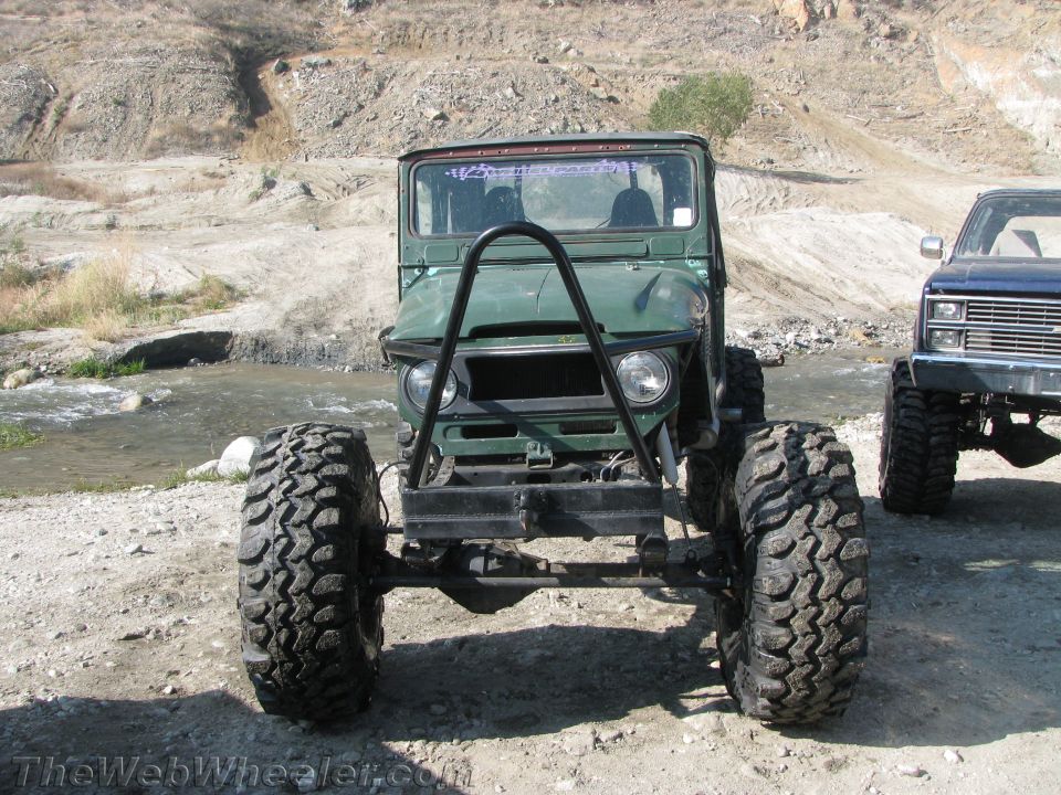 Green off-road vehicle with large tires and a roll cage, parked by a stream next to a blue pickup truck