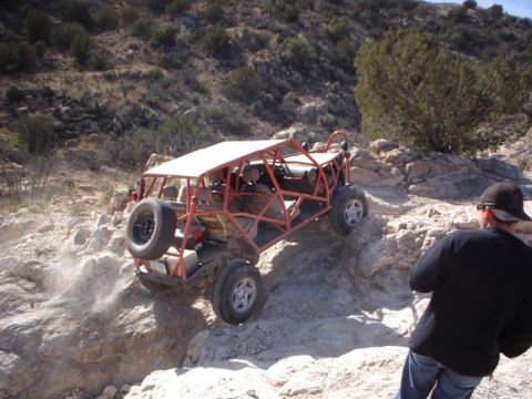 1989 Ford Bronco - An orange dune buggy navigates a challenging rocky trail, kicking up dust as it ascends.