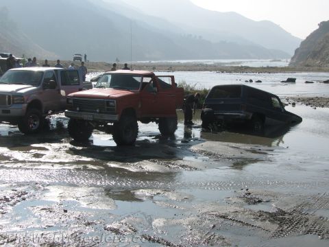 1987 Suzuki Samurai - Several off-road vehicles are stuck in a muddy riverbed, with one red pickup truck partially submerged.