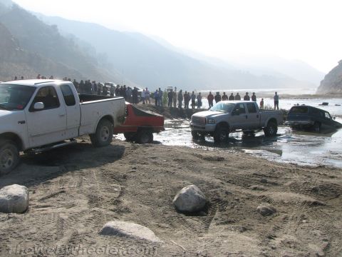 1987 Suzuki Samurai - Multiple vehicles are stuck in the mud near a river with spectators watching.