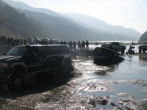 1987 Suzuki Samurai - Several off-road vehicles are stuck in a muddy riverbed, with one truck attempting to tow another out.