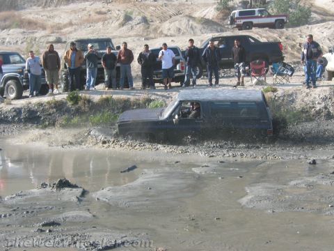 1987 Suzuki Samurai - A dark SUV navigates a muddy obstacle while onlookers observe from the side.