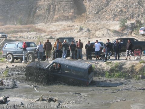 1987 Suzuki Samurai - A group of people watch as a dark blue SUV struggles through a muddy obstacle course.
