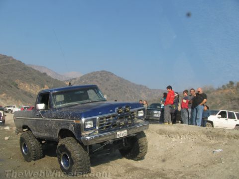 1987 Suzuki Samurai - Modified blue pickup truck with large tires parked on a dirt track with people in the background.