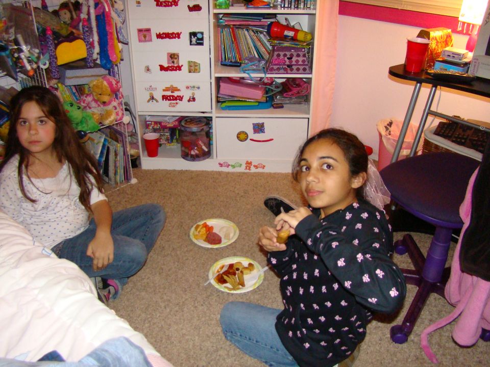 Two girls sit on the floor in a bedroom, eating snacks from paper plates. A shelf unit with books and toys is behind them