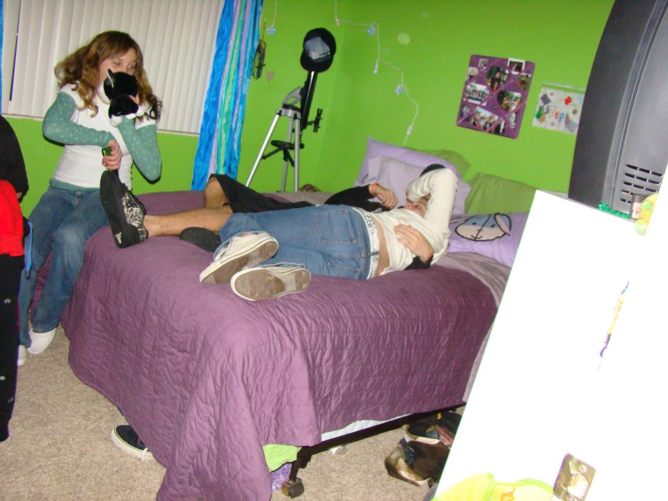 Two teenagers on a purple bed in a green bedroom, one holding a stuffed animal