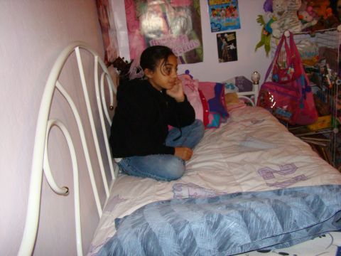 1989 Ford Bronco - A young girl sits cross-legged on a bed in a cluttered bedroom, surrounded by posters and stuffed animals.