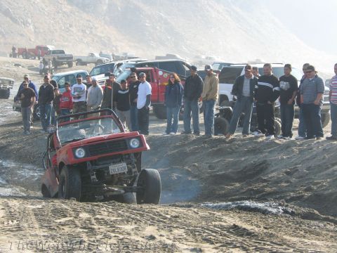 1987 Suzuki Samurai - A group of people watch as a red Suzuki Samurai navigates a sandy incline, kicking up dust.