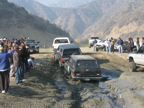 1987 Suzuki Samurai - A group of off-road vehicles tackle a challenging muddy course, observed by a crowd of onlookers.