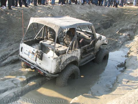 1987 Suzuki Samurai - A Suzuki Samurai is stuck in a muddy obstacle course, with onlookers gathered on the embankment.