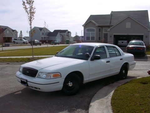 1999 Ford Crown Victoria - A white sedan is parked in front of a suburban house with a garage containing another vehicle.