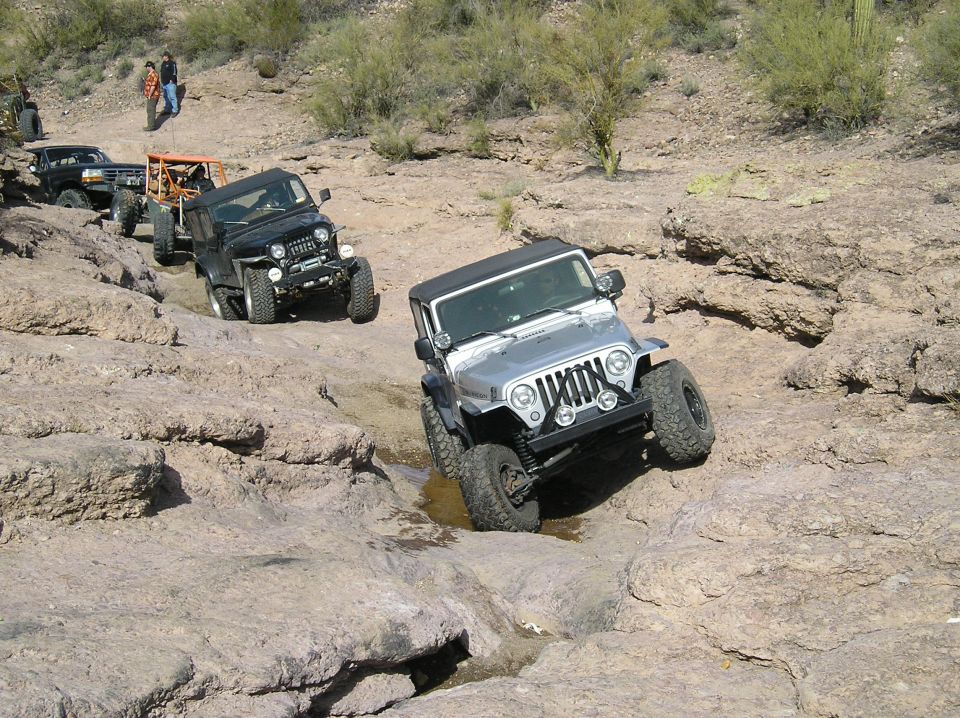 Several Jeeps driving over rocks on a trail. One silver Jeep is in the foreground, with a black Jeep and a truck behind it