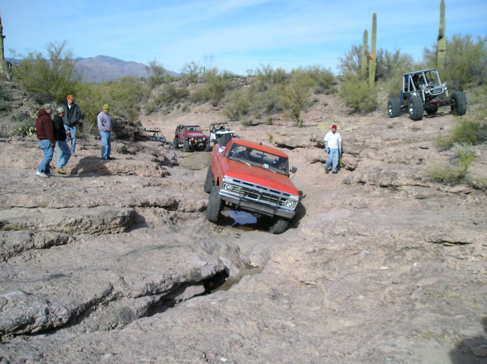 Red pickup truck and other off-road vehicles on a rocky trail with cacti and mountains in the background