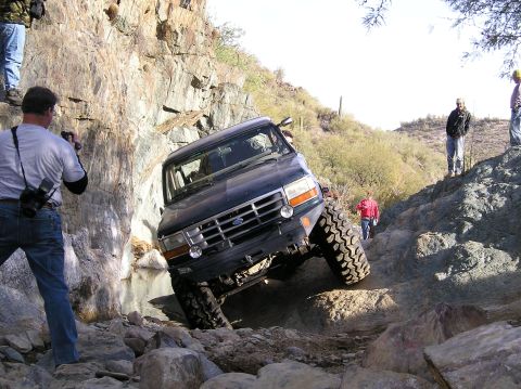 1986 Toyota Hilux - A dark blue Ford F-series truck maneuvers through a challenging rocky terrain with onlookers present.