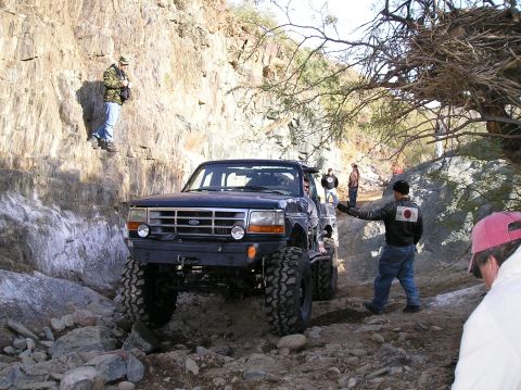 1986 Toyota Hilux - A Ford F-Series truck navigates a rocky off-road trail with several people watching.