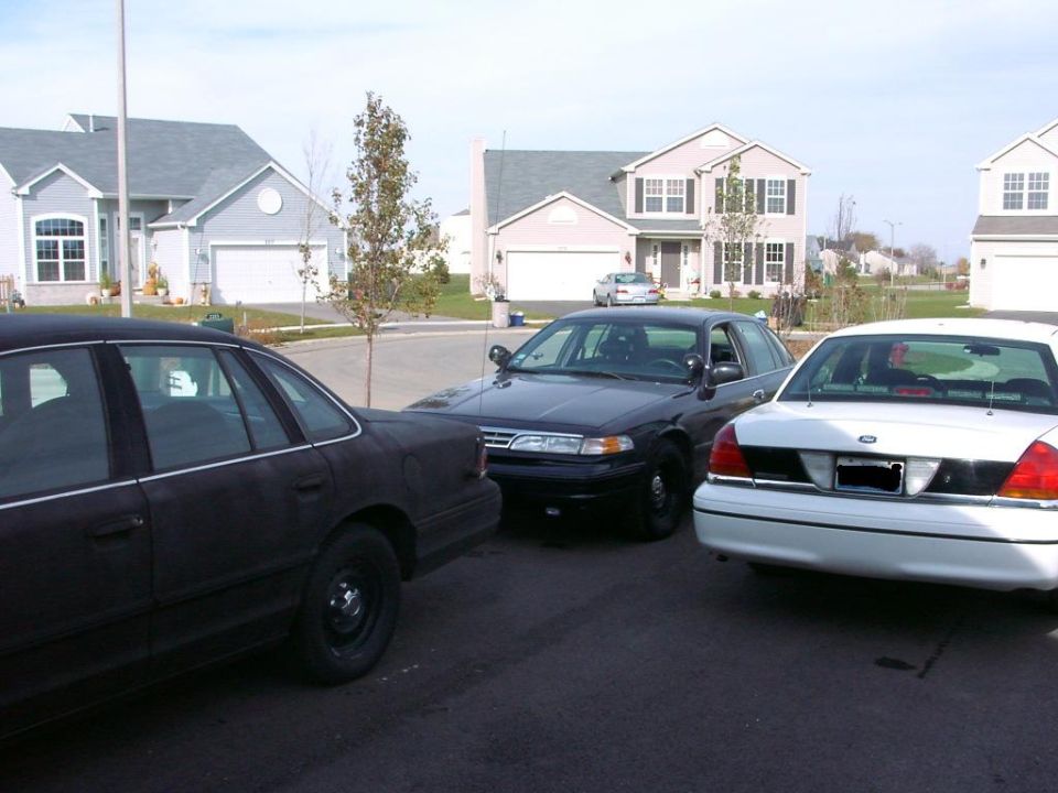 Three Ford Crown Victorias parked in a suburban driveway. One is black, one is dark blue, and one is white
