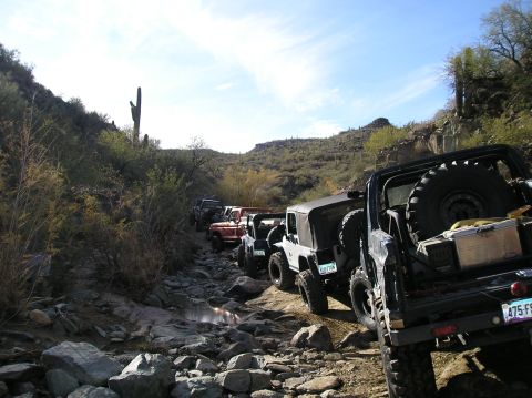 1986 Toyota Hilux - Several off-road vehicles are lined up on a rough, rocky trail in a desert landscape under a clear blue sky.