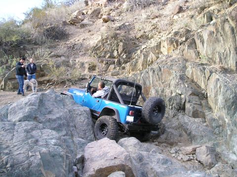 1986 Toyota Hilux - A blue Jeep Wrangler tackles a challenging rocky terrain, with onlookers observing its progress.
