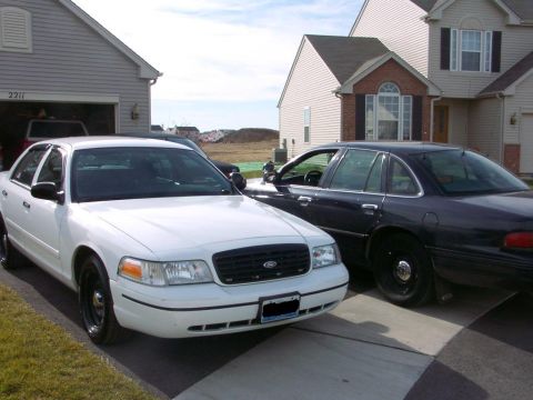 1999 Ford Crown Victoria - Two Ford Crown Victorias, one white and one dark colored, are parked in a driveway in front of houses.