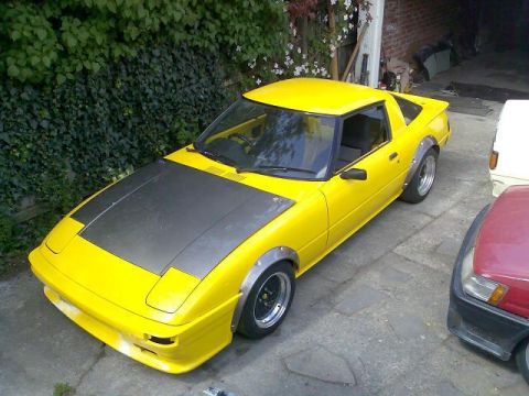 1983 Mazda RX-7 - A yellow 1983 Mazda RX-7 is parked outdoors next to other vehicles.
