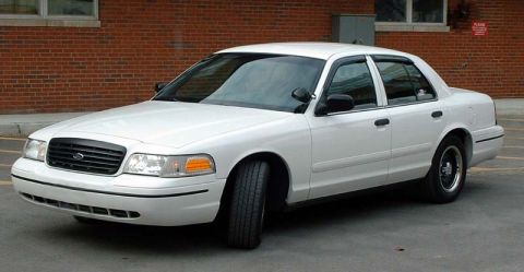 1999 Ford Crown Victoria - A white sedan is parked outside a brick building. The front of the car is angled towards the camera.