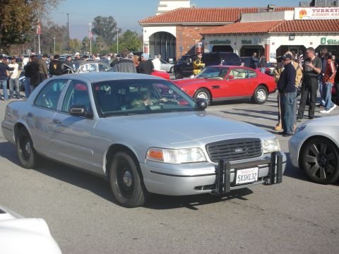 1998 Ford Crown Victoria - A silver 1998 Ford Crown Victoria police car is parked at a car meet, with other vehicles and people in the background.
