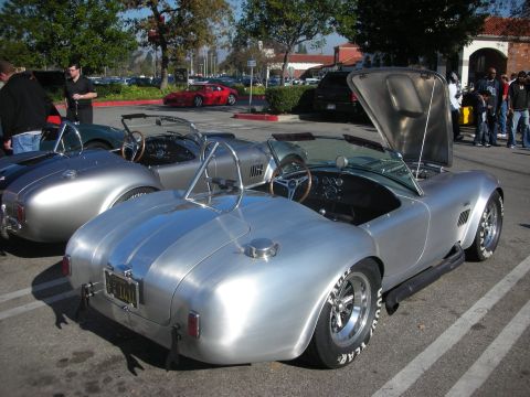 1998 Ford Crown Victoria - A pair of silver AC Cobra replicas are parked side-by-side at a car meet, one with its hood open.