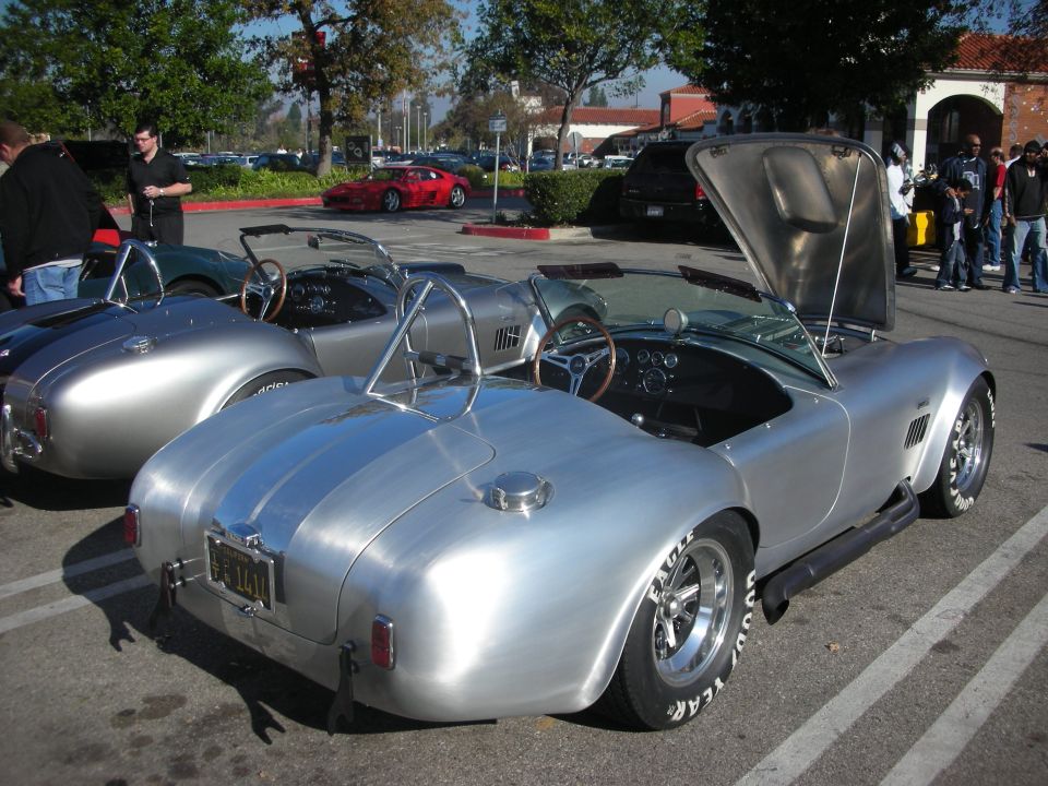 Rear three-quarter view of silver Cobra roadsters with trunk open at a car show