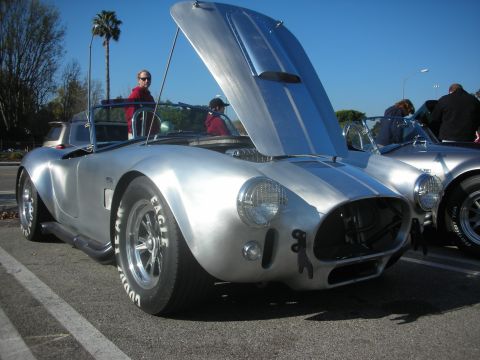 1998 Ford Crown Victoria - A silver AC Cobra with racing stripes and white letter tires is parked with its hood open, alongside another similar vehicle.