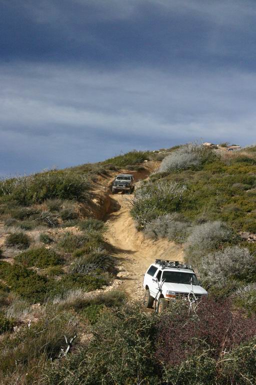 White SUV with roof rack climbing a dirt trail, another vehicle behind it
