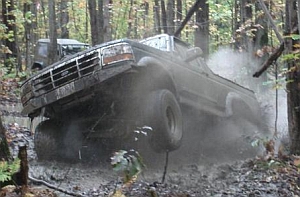 1986 Ford Bronco - A dark Ford Bronco maneuvers through a challenging muddy off-road course, with another vehicle visible in the background.