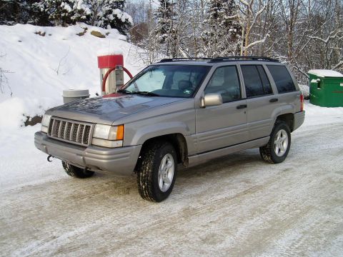 2000 Ford E350 - A silver Jeep Grand Cherokee is parked on a snow-covered road with trees and a dumpster in the background.