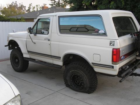 1990 Ford Bronco - White 1990 Ford Bronco parked on a concrete driveway, showing off-road modifications and a flat rear tire.