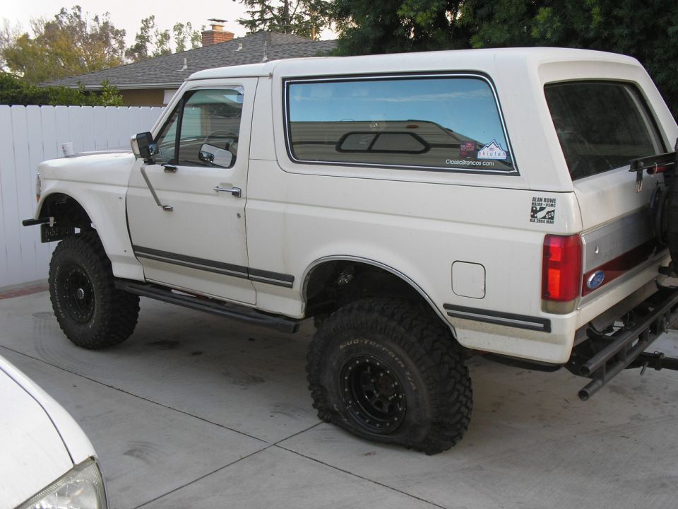 White 1990 Ford Bronco with large off-road tires and a flat rear tire
