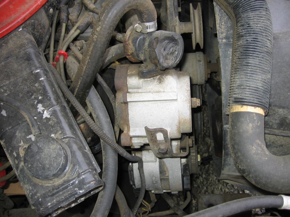 Close-up of an engine bay showing an alternator, various hoses, and wiring. Some parts appear rusty