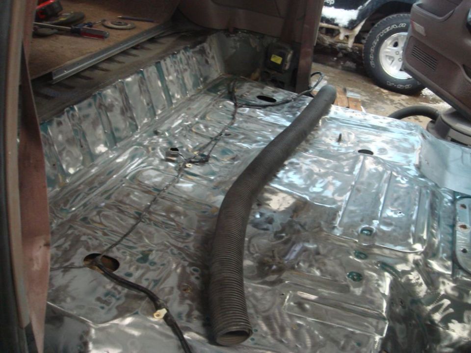 Interior floor of a vehicle stripped down to bare metal, with a black hose running across it. Another vehicle is visible outside