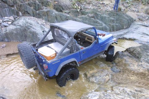 1994 Ford Bronco - A blue Jeep Wrangler is shown off-roading through a rocky stream, with a spare tire mounted on the back.