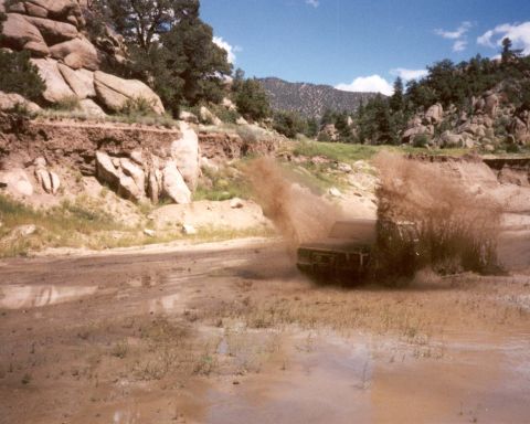 1979 Ford Bronco - A dark 1978 Ford Bronco navigates a muddy off-road course, creating a large spray of dirt and water.