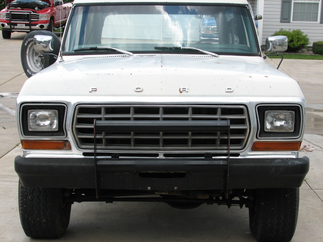 Front view of a white Ford Bronco with a spare tire mounted on the driver's side fender. A red pickup truck is visible in the background