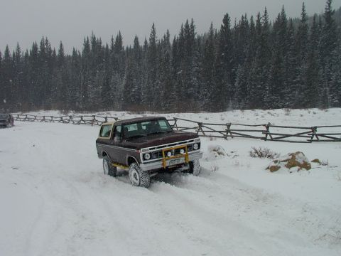 1979 Ford Bronco - A maroon Ford Bronco navigates a snowy landscape with a wooden fence and pine trees in the background.