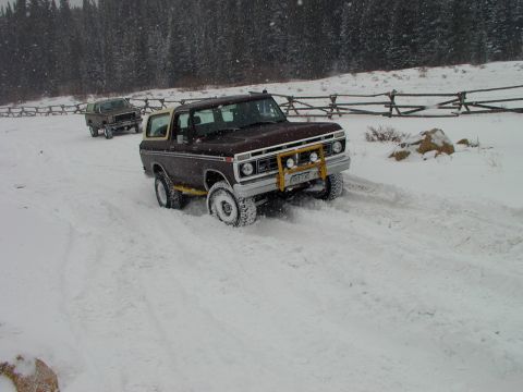 1979 Ford Bronco - Two SUVs navigate a snowy path with trees and a wooden fence in the background.