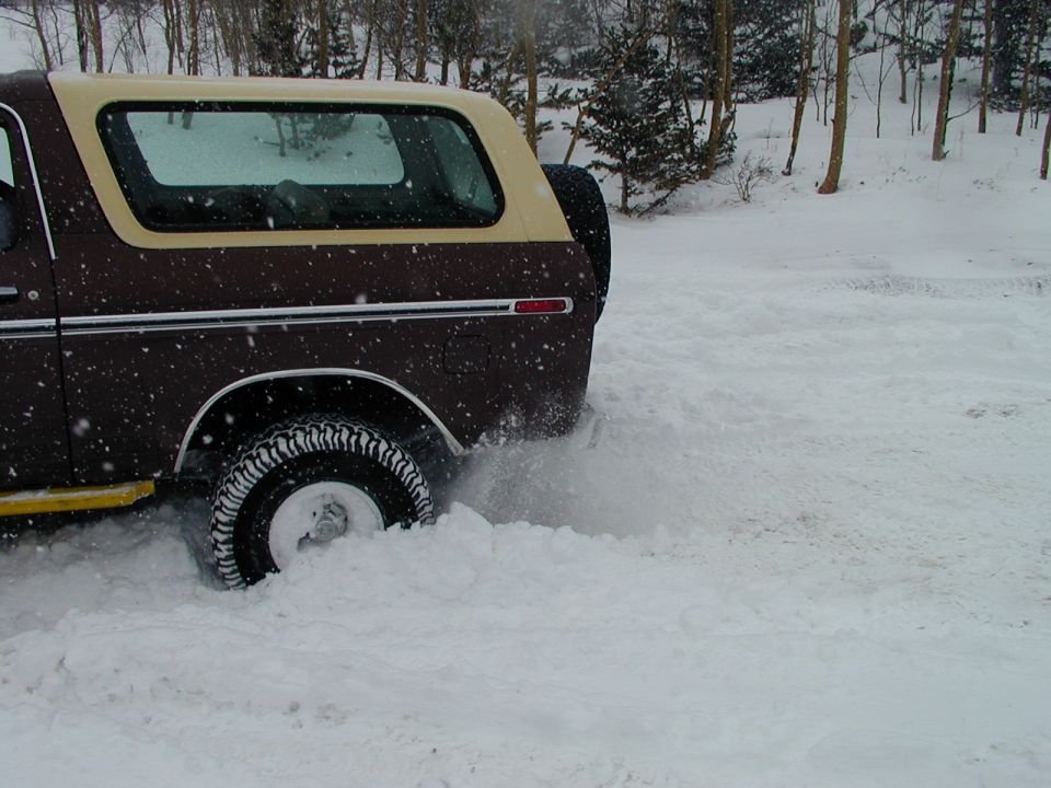 1978 Ford Bronco rear quarter panel and tire kicking up snow. Tan hardtop