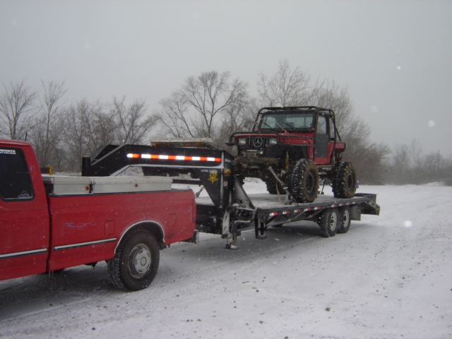 Red off-road vehicle with a roll cage on a flatbed trailer being towed by a red pickup truck in the snow