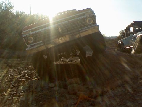 1974 Ford F-250 - A red Ford truck navigates a rough, rocky terrain under a bright sun, with another vehicle in the background.