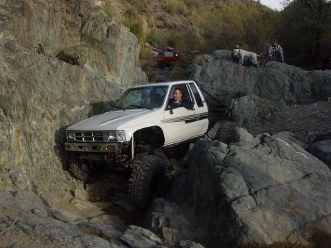 1986 Toyota Hilux - A modified pickup truck tackles a challenging rocky terrain, with another vehicle and people observing in the background.